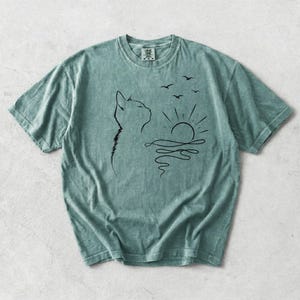 May include: A sage green short-sleeve t-shirt featuring a black line art graphic on the chest. The design depicts the profile of a cat looking towards a stylised sun rising over wavy water, with three small birds flying in the sky above the sun.