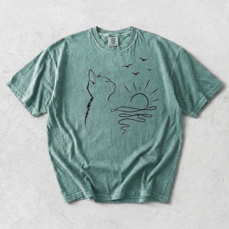 May include: A sage green short-sleeve t-shirt featuring a black line art graphic on the chest. The design depicts the profile of a cat looking towards a stylised sun rising over wavy water, with three small birds flying in the sky above the sun.