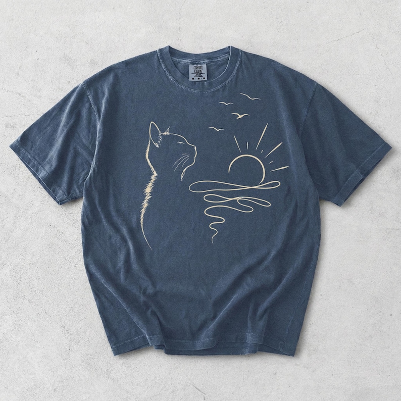 May include: A dark blue short-sleeved t-shirt featuring a light-coloured line art graphic of a cat in profile looking towards a rising sun over water with three birds flying above. The shirt has a crew neckline and a faded, washed-out fabric texture.