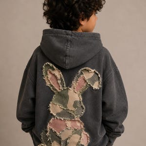 May include: A charcoal gray hooded sweatshirt featuring a large, appliquéd rabbit silhouette on the back. The rabbit is constructed from patches of camouflage fabric in shades of green, brown, and muted pink, with frayed, raw edges throughout.