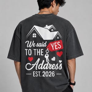 May include: A charcoal gray short-sleeve t-shirt featuring a white graphic of a house on the back. The text reads We said YES TO THE Address EST. 2026, accompanied by a red tag, a silver key, and several red and white heart icons.