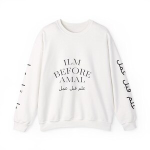 May include: A white sweatshirt with the text "ILM BEFORE AMAL" in black, along with Arabic script. The sleeves also feature black text. The sweatshirt has a crew neck and a relaxed fit.