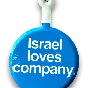 Könnte beinhalten: Ein blauer Knopf mit dem weißen Text "Israel loves company". Der Knopf hat einen weißen Stiel und eine kreisförmige Oberseite. Der Knopf ist hellblau.