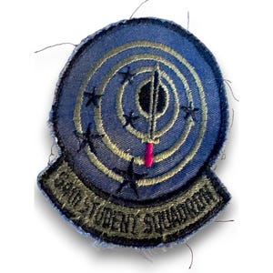 Pode incluir: Um emblema bordado circular com fundo azul apresentando um ícone de espada central, círculos concêntricos e cinco estrelas pretas. A aba inferior é verde-oliva com o texto 64th STUDENT SQUADRON bordado em fio preto.