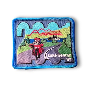 Könnte beinhalten: Gestickter Aufnäher mit einem roten Motorrad mit zwei Fahrern auf einer kurvenreichen Straße. Der Aufnäher enthält den Text "Americade" und "Lake George NY" mit einem blauen Rand und einem landschaftlichen Hintergrund.