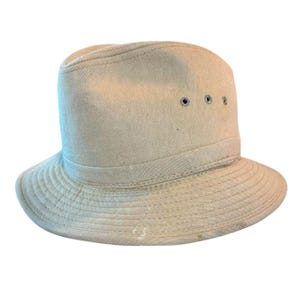 Op de afbeelding: Een beige hoed met een gestructureerde kroon en een brede, licht gebogen rand. De hoed heeft een band rond de basis van de kroon en drie kleine, gelijkmatig verdeelde oogjes. De rand heeft een reeks gestikte lijnen.