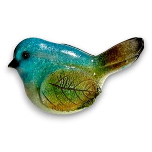May include: A ceramic bird figurine with a glossy finish. The bird features a teal head and back, transitioning to green and brown on the body and tail. A leaf imprint is visible on the side. The bird has a black beak and a white background.