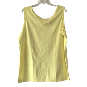 Puede incluir: Una camiseta sin mangas de color amarillo pálido con cuello redondo y un delicado detalle de encaje en el escote. La camiseta sin mangas está hecha de una tela suave y ligera y cuelga de una percha blanca sobre un fondo blanco.