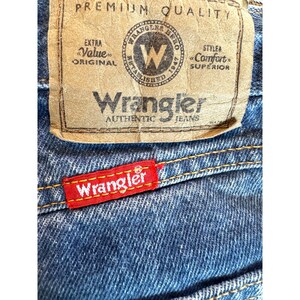 May include: Close-up of blue denim jeans with a red and white Wrangler tag. The jeans have a brown leather patch with the Wrangler logo and text. The jeans are made of a durable denim material.