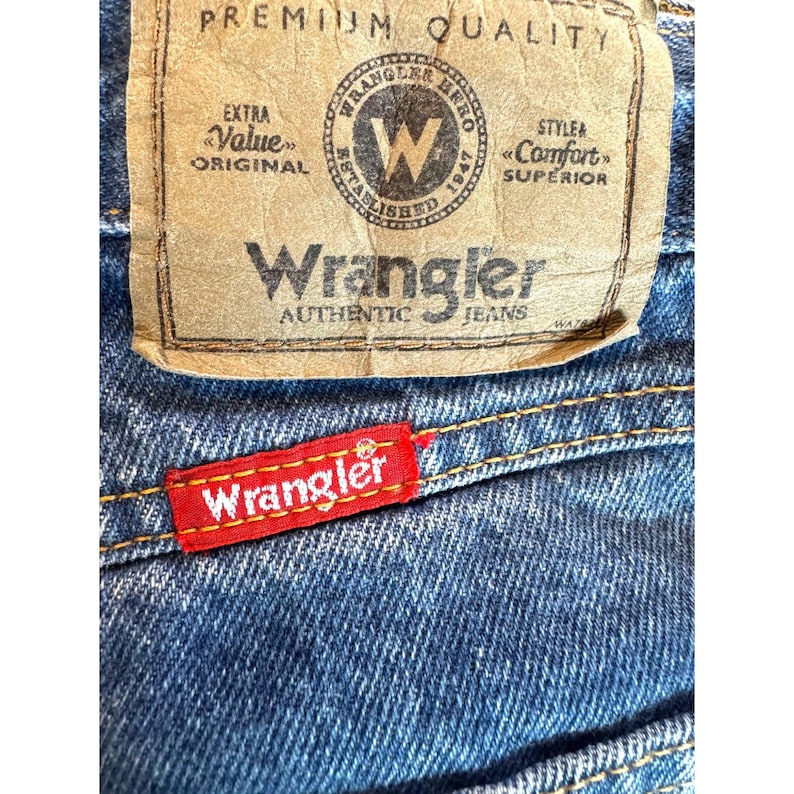 May include: Close-up of blue denim jeans with a red and white Wrangler tag. The jeans have a brown leather patch with the Wrangler logo and text. The jeans are made of a durable denim material.
