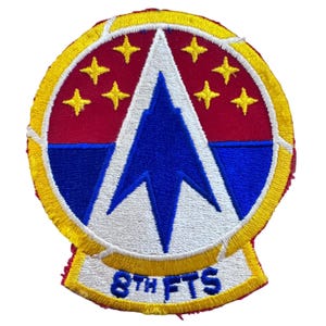 May include: Embroidered patch with a circular design featuring a red background with yellow stars and a blue and white central emblem. The text "8th FTS" is displayed at the bottom. The patch has a yellow border.