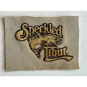 May include: Embroidered patch with the words "Speckled Trout" in a stylized font. The patch features a detailed illustration of a trout in shades of gold and brown, set against a beige background.