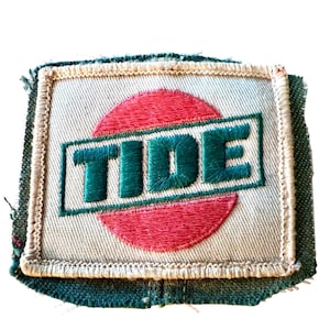 Op de afbeelding: Een rechthoekige geborduurde patch met een beige achtergrond en een rood cirkelvormig ontwerp in het midden. Het woord TIDE is in donkergroene draad in het midden geborduurd in een schuin geplaatst rechthoekig kader met een witte rand.