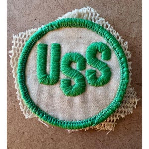 May include: Embroidered patch with the letters "USS" in bright green, encircled by a green border. The patch is on a beige fabric background, with a cream-colored, open-weave fabric extending from the edges. The patch is likely a military or nautical emblem.