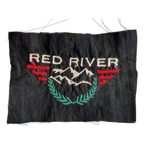 Peut inclure: Écusson brodé noir avec l'inscription blanche « RED RIVER », surmontant une chaîne de montagnes et une couronne de laurier verte. Des accents rouges et blancs sont présents. Le patch est rectangulaire.