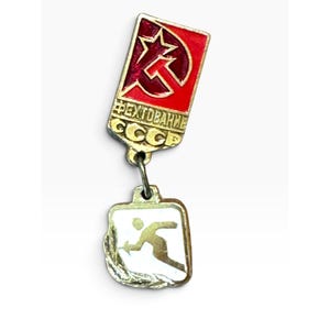 Könnte beinhalten: Eine zweiteilige Anstecknadel mit einem rechteckigen Oberteil auf rotem Emaille-Hintergrund, einem goldenen Stern- und Sichel-Emblem sowie dem kyrillischen Text "ФЕХТОВАНИЕ СССР". Darunter hängt ein kleinerer, quadratischer Anhänger mit einem Fechter.