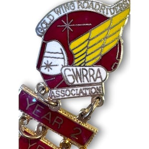 May include: A metal pin with the words "GOLD WING ROADRIDERS" in a curved banner. The pin features a red and yellow wing design, the letters "GWRRA ASSOCIATION", and a banner that says "YEAR 2".