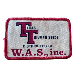May include: A rectangular embroidered patch with a cream-colored background and a red stitched border. It features the maroon text "TEXAS TRIUMPH SEEDS" and "DISTRIBUTED BY W.A.S., inc." with a large maroon "TT" logo on the left side of the patch.