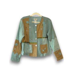 Puede incluir: Chaqueta patchwork azul claro y marrón con bordados florales y cierre de botones. La chaqueta presenta un cuello, dos bolsillos delanteros y costuras decorativas. La chaqueta de manga larga está colgada en una percha de madera.