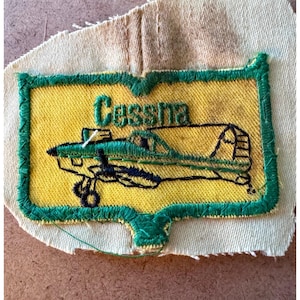 May include: Embroidered patch featuring a yellow background with a green border. The patch displays a green and black airplane with the name "Cessna" in green lettering. The patch is sewn onto a beige fabric backing.
