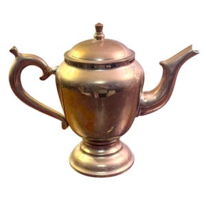 May include: A metallic teapot with a polished copper-toned finish, featuring a rounded body, a curved handle, a tapered spout, and a matching lid with a small finial knob. The teapot stands on a circular pedestal base with a smooth surface.