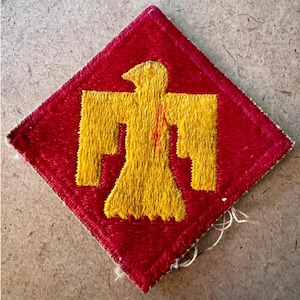 Op de afbeelding: Een rood en geel geborduurde patch met een geel vogel-embleem op een rode ruit. De vogel heeft uitgespreide vleugels. De patch is waarschijnlijk vintage en vertoont tekenen van slijtage.