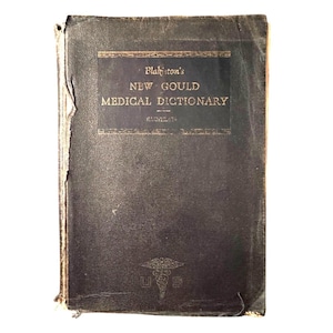 Puede incluir: Un diccionario médico antiguo con una cubierta oscura. El título "Blakiston's NEW GOULD MEDICAL DICTIONARY" está impreso en letras doradas. El libro muestra signos de desgaste, lo que indica edad y uso. El símbolo médico estadounidense está en la parte inferior.