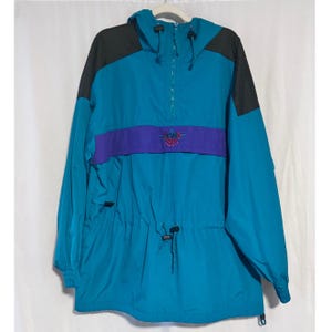 May include: Teal, purple, and black anorak jacket with a hood and a half-zip. The jacket has a purple band across the chest with a logo. It features a drawstring at the waist and is made of a lightweight material.