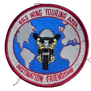 May include: Embroidered patch with a motorcycle design. The patch features a motorcycle in the center, set against a blue background with a world map. The text "GOLD WING TOURING ASSN." and "DESTINATION FRIENDSHIP" are in a circular pattern.