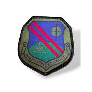 May include: A shield-shaped embroidered patch with a black border. The design features a blue field, a green hill with a gold triangle pattern, two diagonal red stripes, a sword, a target symbol, and a banner reading 4461ST SUPPORT GROUP.