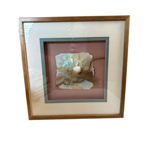 Può includere: Un'opera d'arte da parete quadrata incorniciata con uno sfondo ad acquerello azzurro e rosa, juta a strati, conchiglie bianche e piccoli frammenti di corallo bianco. L'opera è racchiusa in una cornice di legno con un bordo bianco e blu.