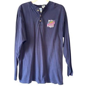 Op de afbeelding: Een marineblauw henley-shirt met lange mouwen en een knoopsluiting met drie knopen bij de halslijn. Op de linkerborst bevindt zich een embleem met de tekst 2005 Wrangler National Finals Rodeo Las Vegas, voorzien van rode, witte en blauwe grafische elementen.