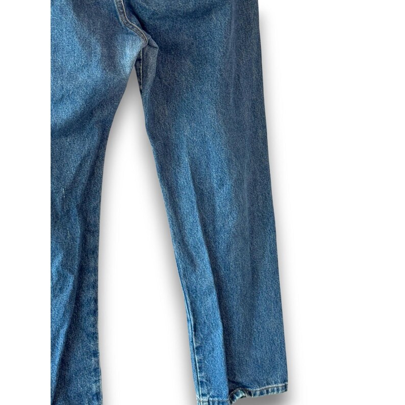 May include: Close-up of blue denim jeans. The jeans have a classic, straight-leg fit and a medium wash. The fabric shows subtle fading and wear, suggesting a vintage or well-loved appearance. The jeans are unhemmed.