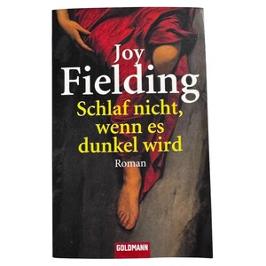 May include: A book cover titled Joy Fielding Schlaf nicht, wenn es dunkel wird Roman. The cover features a dark background with a person wearing a red garment, showing a bare foot and a hand. The Goldmann logo is at the bottom in red.