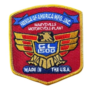 May include: Embroidered patch with a red background and a gold border. The patch features a blue banner with "HONDA OF AMERICA MFG. INC." and "MARYSVILLE MOTORCYCLE PLANT." A gold eagle design with "GL 1500" is in the center, with "MADE IN THE U.S.A." at the bottom.