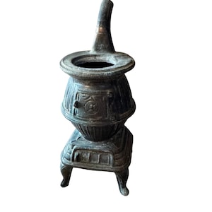 May include: A small, antique cast iron stove with a dark, aged finish. The miniature stove features a round body, a chimney, and decorative details. It stands on four small legs and has a door-like design on the front.