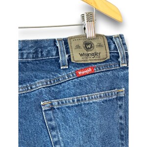 May include: Close-up of blue denim Wrangler jeans. The jeans are hanging on a wooden hanger. The Wrangler label is visible on the back pocket, with the brand name in red. The jeans have a classic five-pocket design.