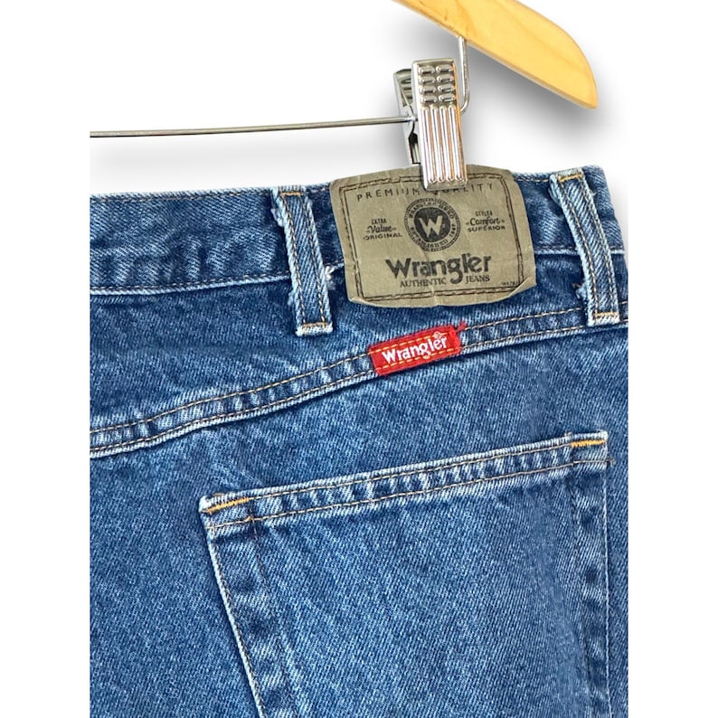 May include: Close-up of blue denim Wrangler jeans. The jeans are hanging on a wooden hanger. The Wrangler label is visible on the back pocket, with the brand name in red. The jeans have a classic five-pocket design.