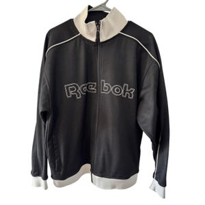 Puede incluir: Chaqueta deportiva negra con cremallera, ribete blanco y el logotipo de Reebok en costuras blancas en el pecho. La chaqueta tiene cuello y puños blancos. La chaqueta está colgada de una percha.