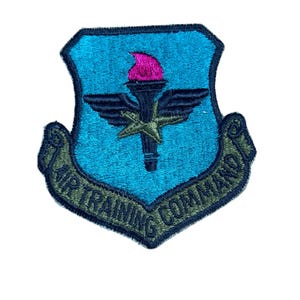 May include: An embroidered shield-shaped patch with a bright blue background. It features a central dark blue torch with a pink flame, black wings, and a green star. A green banner at the bottom displays the text AIR TRAINING COMMAND.