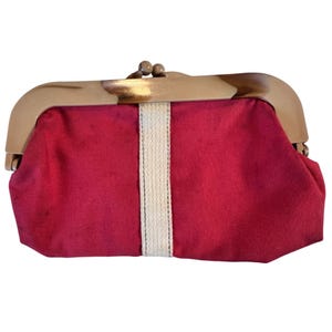 May include: A red clutch with a cream-colored vertical stripe down the center. The frame is a light brown color with a clasp closure. The clutch is a rectangular shape.