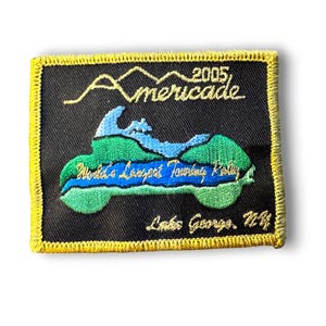 May include: Embroidered patch with a black background and a yellow border. The patch features a green and blue motorcycle design with the text "Americade 2005" and "World's Largest Touring Rally". The text "Lakes George, NY" is also included.
