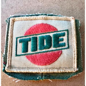 Könnte beinhalten: Gestickter Aufnäher mit dem Wort "TIDE" in grünen Blockbuchstaben auf einem roten Kreis. Der Aufnäher hat einen beigen Hintergrund und einen grünen Rand mit ausgefranstem Rand.