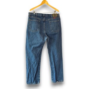May include: Blue denim jeans hanging on a wooden hanger. The jeans have a classic five-pocket design, a leather patch, and a straight leg. The jeans are a medium wash and appear to be in good condition.