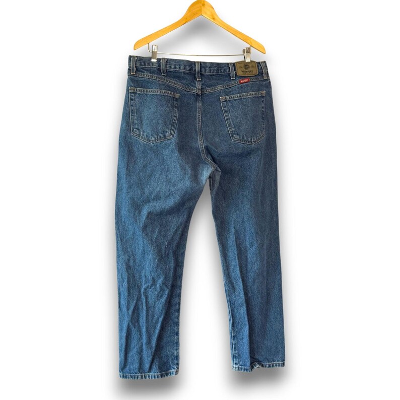 May include: Blue denim jeans hanging on a wooden hanger. The jeans have a classic five-pocket design, a leather patch, and a straight leg. The jeans are a medium wash and appear to be in good condition.