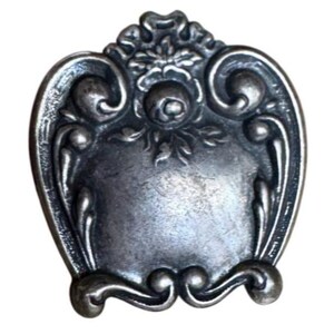 Könnte beinhalten: Ein schildförmiger Metallknopf mit dunkel-silbernem Finish. Die Oberfläche zeigt ein geprägtes Blumenmotiv in der oberen Mitte, umgeben von komplexen Schnörkeln und geschwungenen Zierrändern, die einen glatten, schlichten Mittelbereich einrahmen.