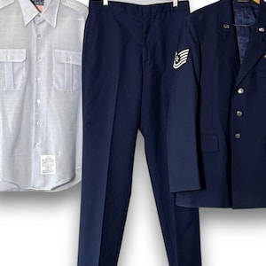Peut inclure: Une chemise boutonnée gris clair avec deux poches de poitrine, un pantalon bleu marine et un blazer bleu marine avec des boutons argentés et un écusson. Les vêtements sont suspendus à des cintres sur fond blanc.