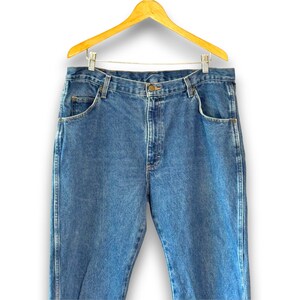 May include: Blue denim jeans hanging on a wooden hanger. The jeans have a classic five-pocket design with a button and zipper closure. The denim has a medium wash with subtle fading.