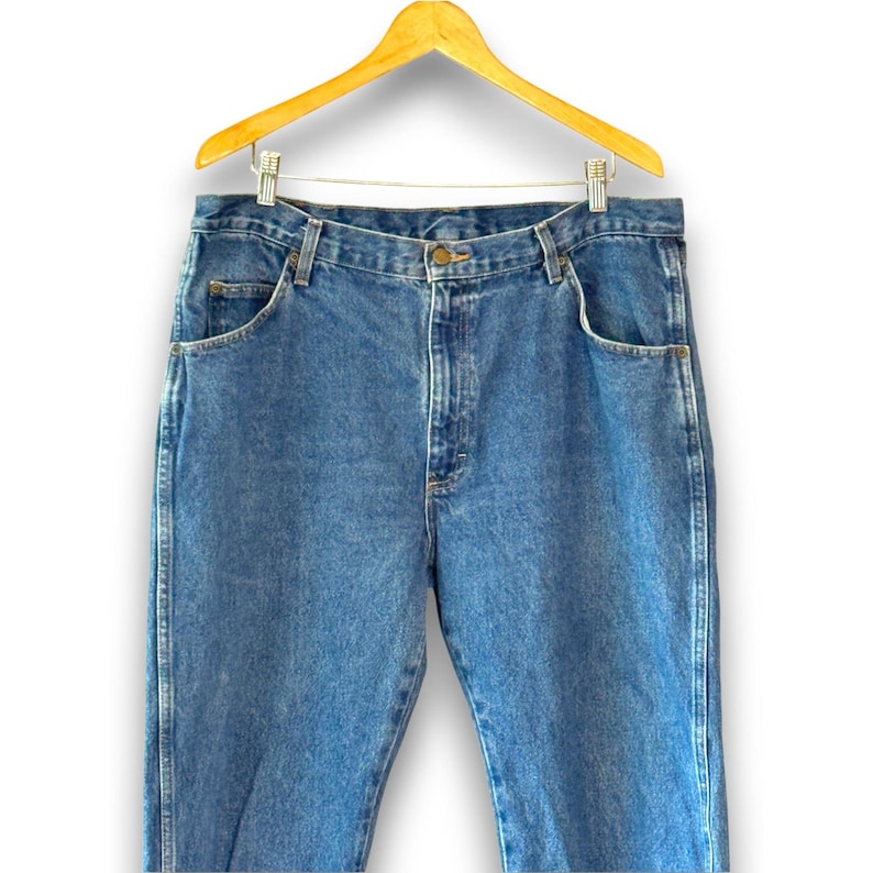 May include: Blue denim jeans hanging on a wooden hanger. The jeans have a classic five-pocket design with a button and zipper closure. The denim has a medium wash with subtle fading.