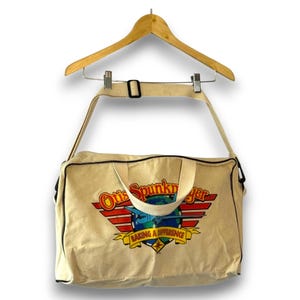 May include: Beige canvas messenger bag with black trim, a white handle, and a long adjustable strap. The bag features a colorful logo with the text "Otis Spunkmeyer" and "Making a Difference" on the front.